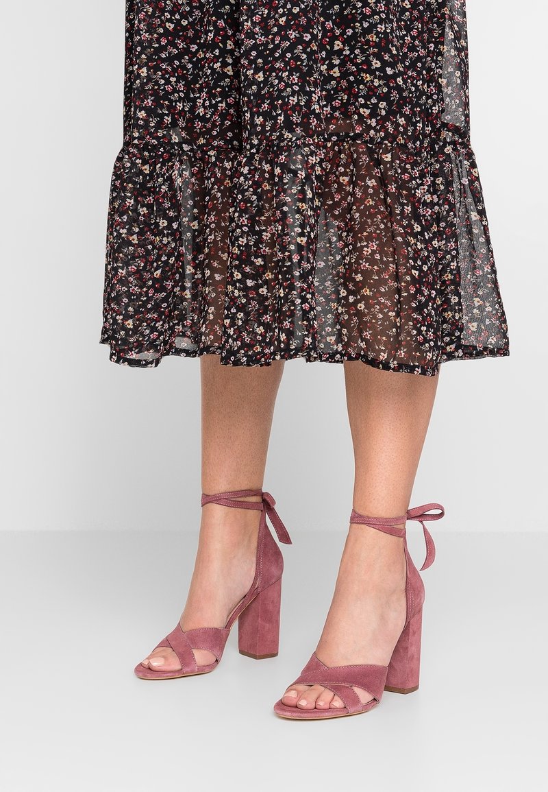 Talons hauts ouverts en suede rose avec un talon bloc, des liens à la cheville et des sangles croisées. Portés avec une robe noire à fleurs.
