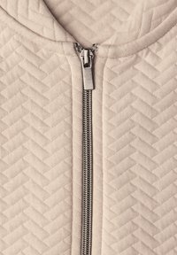 Béžová prošitá mikina s kapucí s texturovaným vzorem rybí kosti, vybavená stříbrným zipem a podšitou kapucí. Zip vede po celé délce přední části.