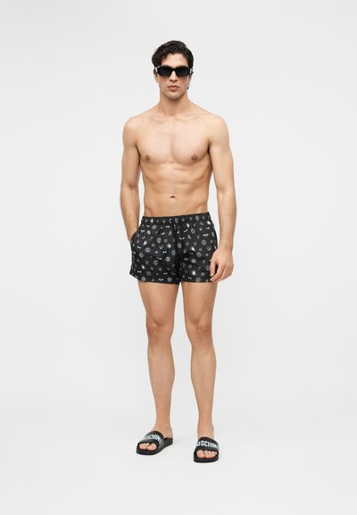 Skjorteløs mann iført svarte mønstrede badeshorts, svarte solbriller og svarte sandaler med hvit merkevaretekst, stående foran en ensfarget bakgrunn.