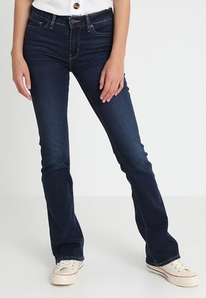 Personne portant un jean slim bootcut bleu foncé et des baskets blanches, debout avec une main posée sur la cuisse devant un fond uni.