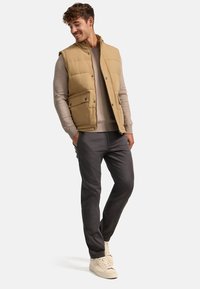 Veste beige rembourrée sur un pull marron clair, associée à un pantalon sombre et des baskets claires. La veste a des poches avant et des boutons-pression.