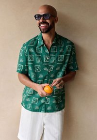 Green short-sleeve shirt with white botanical prints, paired with white linen shorts. Holding an orange, stands against a neutral background.
