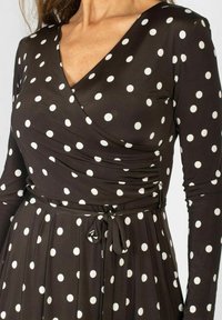 Sukienka z długim rękawem w ciemnym brązie z dużymi białymi kropkami. Posiada dopasowaną górę, marszczone detale oraz pasek do związania w talii.
