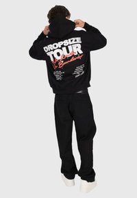 Personne portant un sweat à capuche noir avec "Dropsize Tour No Limits No Boundaries" et des noms de villes imprimés au dos, associé à un pantalon noir et des chaussures blanches.