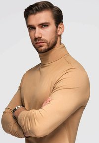 Beige Rollkragensweater, tailliertes Design, glatte Textur, lange Ärmel, minimalistische Nähte, umgeschlagene Bündchen. Modell mit verschränkten Armen.
