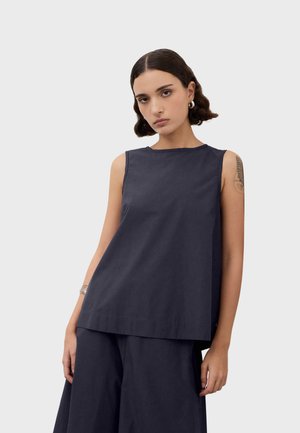 Woman with short dark hair wears sleeveless navy top and matching wide-leg pants, standing against a plain light background.