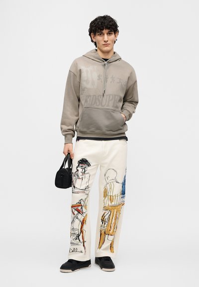 Φούτερ με κουκούλα σε απαλή γκρι απόχρωση με κεντρικό λογότυπο, συνδυασμένο με εκρού παντελόνι που διαθέτει πολύχρωμα καλλιτεχνικά σχέδια και μαύρη τσάντα ώμου.
