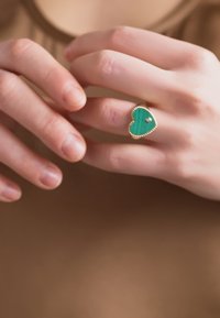 Χρυσό δαχτυλίδι με καρδιοειδή λίθο μαλαχίτη, υφασμένα άκρα και ένα μικρό διακοσμητικό πετράδι, φορεμένο σε χέρι με μπεζ φόντο.