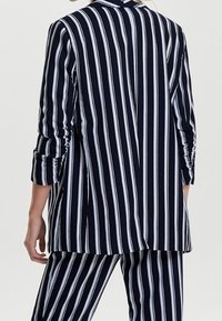 Femme portant un costume bleu marine à rayures blanches verticales, composé d'une veste à manches longues et d'un pantalon assorti, vue de dos.