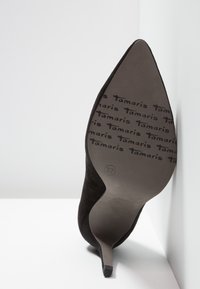 Chaussure à talon haut en suède noir à bout pointu, posée sur le talon, montrant la marque "Tamaris" et la taille 37 sous la semelle.