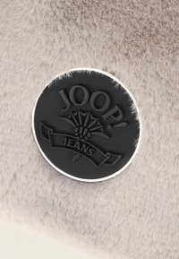 Černá kulatá brož s logem Joop! Jeans na jemném světle béžovém sametovém pozadí.