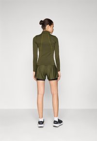Oliven grønn treningstopp med lange ermer og gule sting, kombinert med løse olivenfargede shorts. Modell med svarte joggesko og hvite sokker.