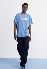 Tricou din bumbac albastru cu logo, asortat cu pantaloni negri. Costumul are mâneci scurte, o croială lejeră și adidași cu accente albastre.