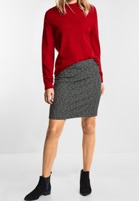 Roter gestrickter Pullover mit Rundhalsausschnitt, kombiniert mit einem Bleistiftrock im schwarz-weißen Hahnentrittmuster. Schwarze Wildleder-Stiefeletten runden das Outfit ab.