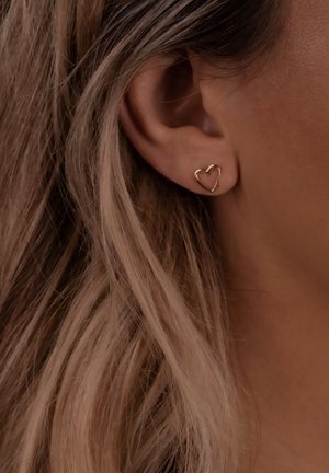 Goldene herzförmige Steckerohrringe mit offenem Design, die an einem durchstochenen Ohr befestigt sind, vor hellbraunem Haar. Einfach und geometrisch.