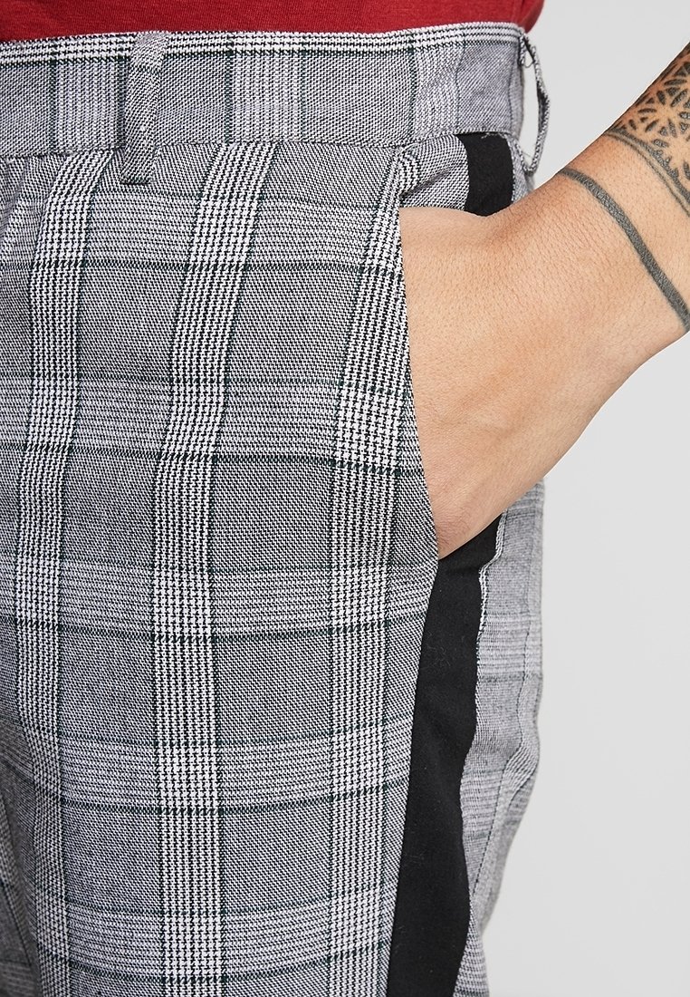 Main avec tatouage reposant dans la poche d’un pantalon gris à carreaux avec une bande noire verticale, associée à un haut rouge.