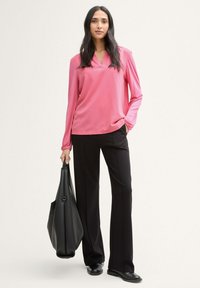 Blusa rosa de manga larga con escote en V, combinada con pantalones amplios negros. Modelo sosteniendo un bolso tote negro texturizado. Textura de tela suave.