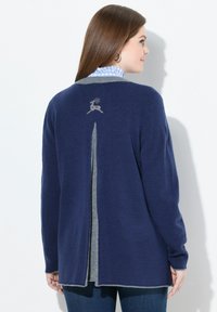 Vrouw in een marineblauwe cardigan met een grijzige verticale streep en een klein rendierpatroon op de bovenrug, over een blauw geruit shirt.