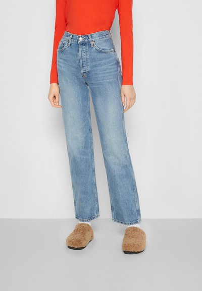 Jeans de ganga de cintura alta, de cor azul clara, com um corte reto, complementados por uma blusa de manga longa sólida em vermelho e uns sapatos de slip-on texturizados em castanho.