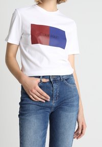 Bílý bavlněný tričko se čtvercovým grafickým prvkem v červené a modré barvě se textem "Calvin Klein" ve středu. Úzký střih, krátké rukávy.