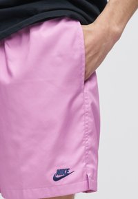 Άτομο που φοράει ανοιχτό μοβ σορτς Nike με το χέρι στην τσέπη, δείχνοντας το ναυτικό μπλε λογότυπο Nike στο κάτω αριστερό πόδι.