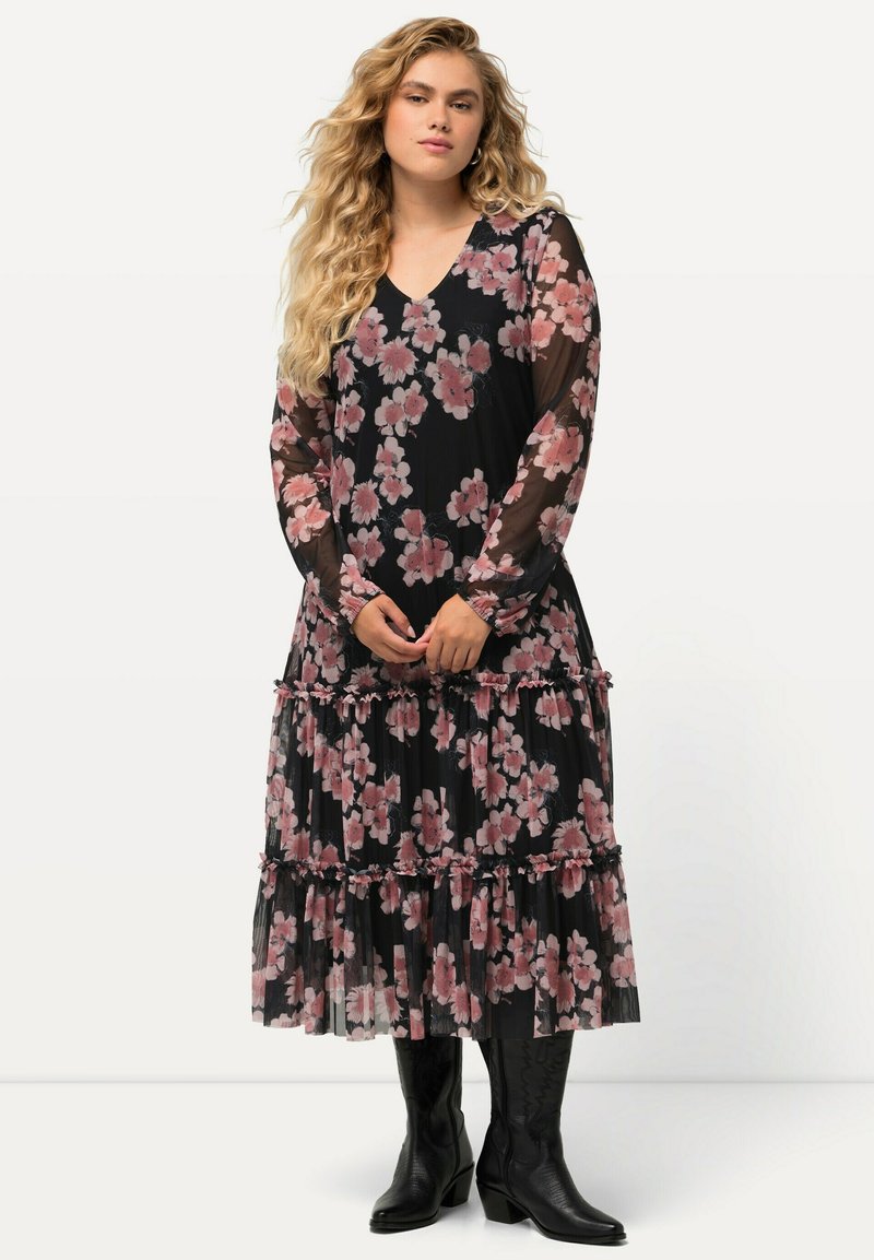 Largo vestido negro con flores rosas, con volantes en capas y mangas largas transparentes. Combinado con botas de vaquero negras. Diseño de escote en V.