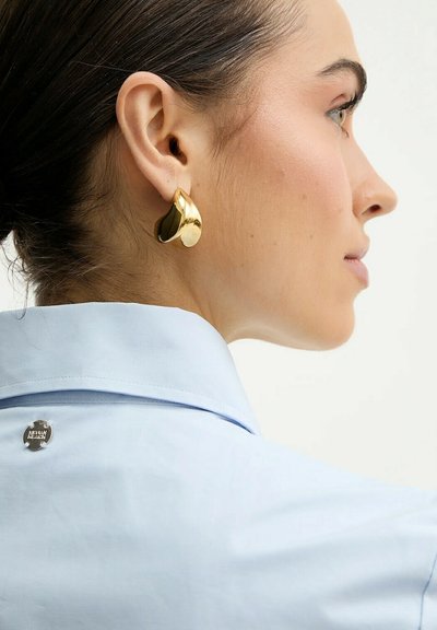 Profilo di una donna che indossa una camicia azzurra con colletto e un grande orecchino d'oro lucido dalla forma irregolare.