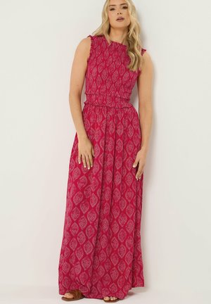 Mujer con vestido rojo sin mangas hasta el suelo con patrones intrincados en rosa claro, posando frente a un fondo blanco liso.