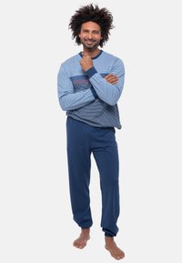 Man with curly hair smiling, wearing blue striped long-sleeve shirt and navy blue pants, standing barefoot against white background.