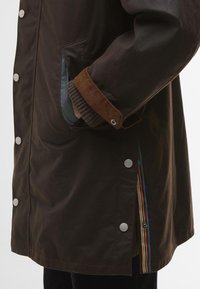 Barna vízálló kabát ezüst patentgombokkal, kontrasztos kordbársony mandzsettákkal, és színes csíkos béléssel, ami az oldalon látható.