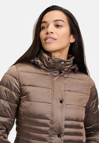 Giacca a vento trapuntata marrone con collo alto, bottoni neri e texture lucida. Presenta una chiusura con zip e cinturini regolabili al collo.