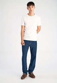 Maglietta bianca di cotone a maniche corte, abbinata a jeans di denim blu scuro e scarpe marroni con lacci, in piedi contro uno sfondo semplice.