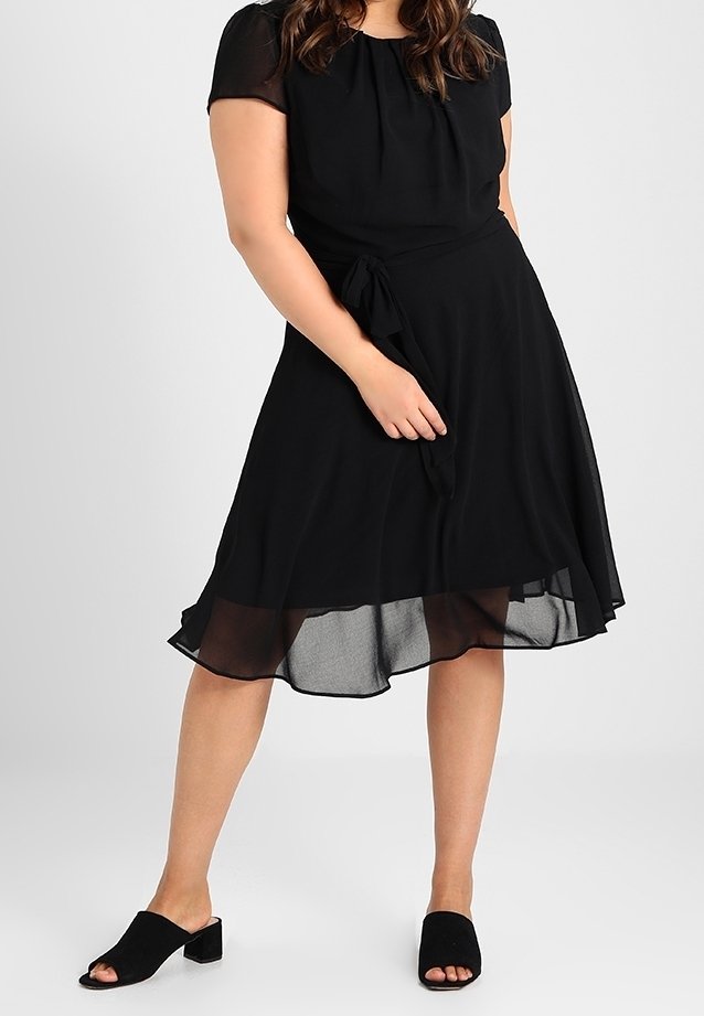 Svart klänning med korta genomskinliga ärmar, åtsittande bodice och en flödande A-linjeformad kjol. Har ett knytband i midjan. Bärs med svarta slip-on skor.