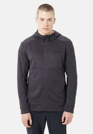 Junger Mann, der eine dunkelgraue Kapuzenjacke mit Reißverschluss trägt und mit entspannten Armen vor einem schlichten hellen Hintergrund steht.