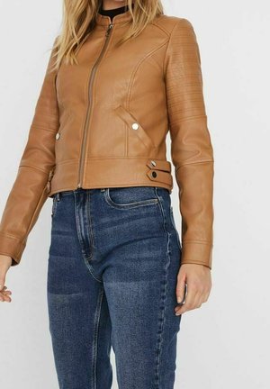 Frau trägt eine hellbraune Lederjacke mit Reißverschluss und Druckknopfdetails und eine hoch taillierte blaue Jeans.