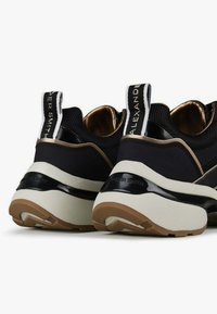 Zwarte sneakers van mesh met glanzende accenten, voorzien van een gouden binnenkant, dikke witte zolen en zwart-witte pull tabs met merknaam.