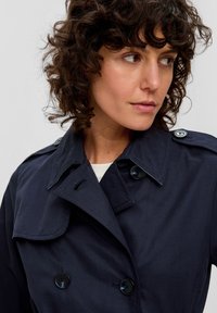 Granatowy płaszcz trench z podwójnym zapięciem, dużym kołnierzem i guzikami; wykonany z gładkiej, teksturowanej tkaniny.