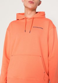 Felpa arancione realizzata in tessuto morbido, con tasca frontale, coulisse e il nome del marchio "SCOTCH & SODA" ricamato in blu.