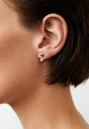 Prim-plan al unei urechi stângi cu păr scurt și închis la culoare, purtând cercei mici aurii în formă dublă de inimă, în formă de picătură.