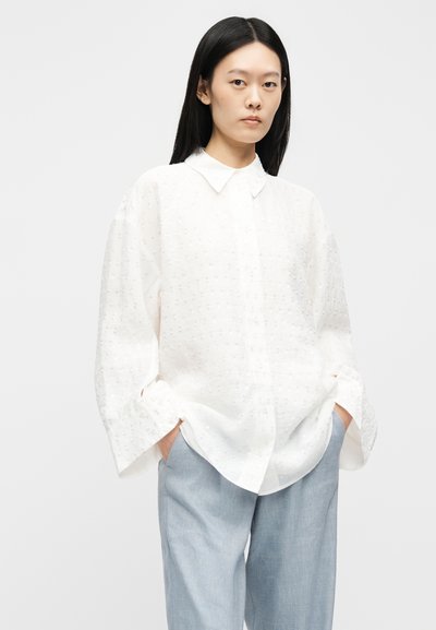 Femeie cu păr negru lung, purtând o bluză albă texturată, largă, cu mâneci largi și pantaloni drepti bleu deschis, mâinile în buzunare.