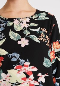 Blusa floreale con sfondo nero, decorata con fiori colorati in rosso, rosa e blu. Comprende foglie verdi e un collo rotondo.