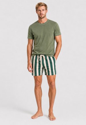 Ein Mann steht barfuß, trägt ein grünes kurzärmeliges T-Shirt und grün-weiß vertikal gestreifte Shorts mit Kordelzug.