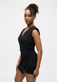 Femme aux cheveux bouclés portant une robe portefeuille noire sans manches, debout devant un fond blanc uni, regardant vers la caméra.