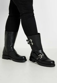 Botas de cuero negras hasta la rodilla con suelas planas. Cuentan con cremallera lateral, detalles de hebilla y una textura suave. El diseño es funcional y resistente.