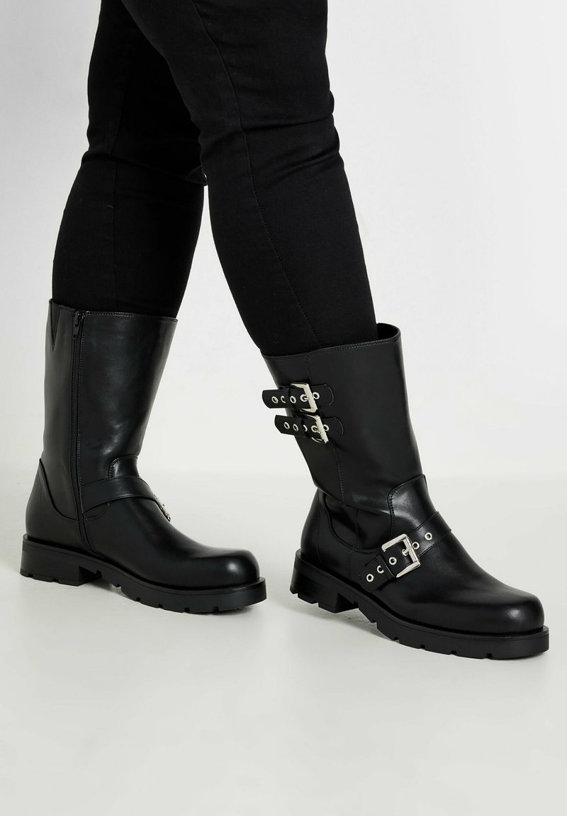 Botas de cuero negras hasta la rodilla con suelas planas. Cuentan con cremallera lateral, detalles de hebilla y una textura suave. El diseño es funcional y resistente.