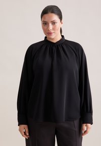 Blusa nera con colletto alto, maniche lunghe e dettagli arricciati sul collo. Presenta polsini abbottonati e una texture di tessuto liscia.