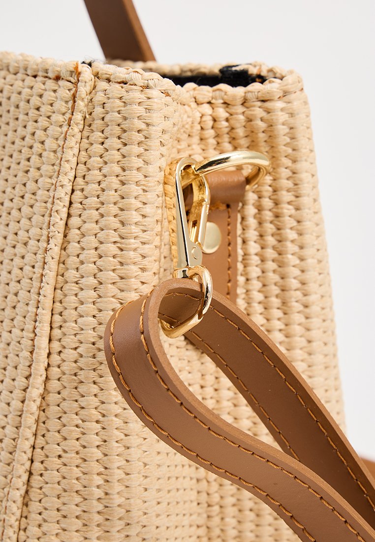 Geweven strohandtas in beige, met een gouden metalen clip en een bruine leren schouderband met stikseldetails, die een gestructureerd oppervlak laat zien.