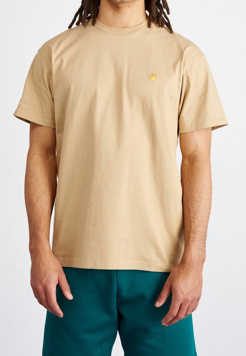 T-shirt in cotone beige con collo rotondo, maniche corte e un piccolo logo giallo sul petto. Texture liscia, vestibilità rilassata.