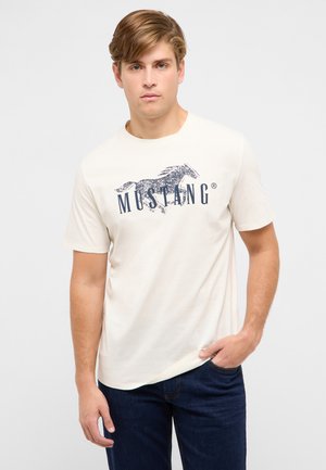 Junger Mann trägt ein weißes T-Shirt mit einem dunkelblauen galoppierenden Pferd und dem Schriftzug "MUSTANG", kombiniert mit dunkelblauen Jeans.