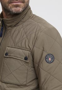 Quilted, olive green jacket with a high collar, zip closure, and two front pockets. Features a round logo patch on the sleeve.